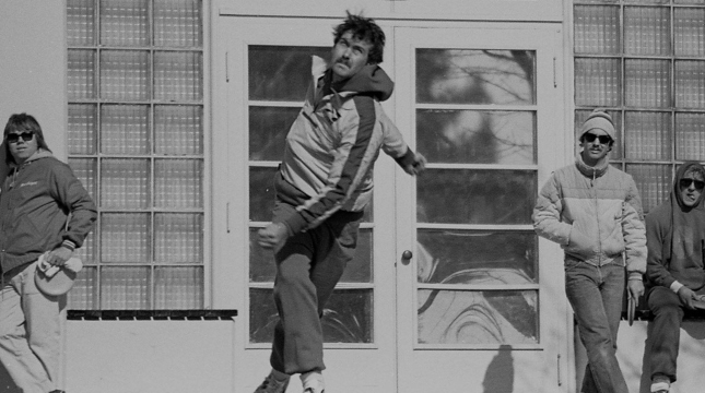 Black and White image of a disc golfer after releasing the disc, with a few players leaning against a building in the background observing. Image appears to be taken in the 1970's.