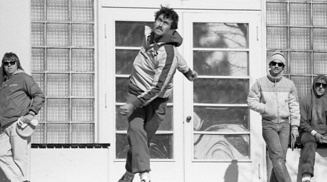 Black and White image of a disc golfer after releasing the disc, with a few players leaning against a building in the background observing. Image appears to be taken in the 1970's.